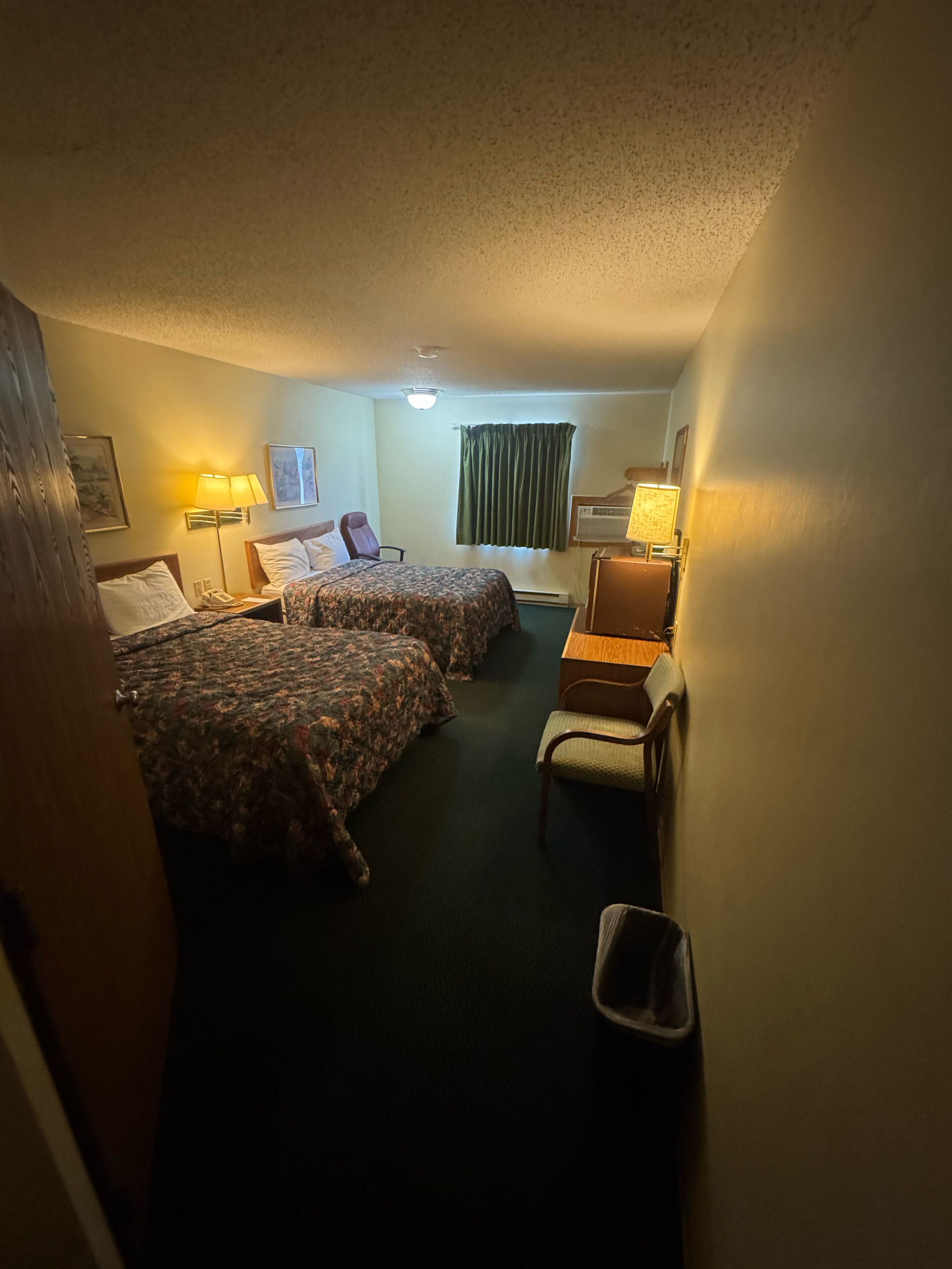 A hotel room with two beds, a desk, a chair, a lamp, and a trash bin.