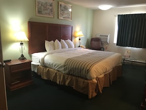 A hotel room with a large bed, two bedside lamps, a desk with a telephone, an air conditioning unit, and a window with curtains.