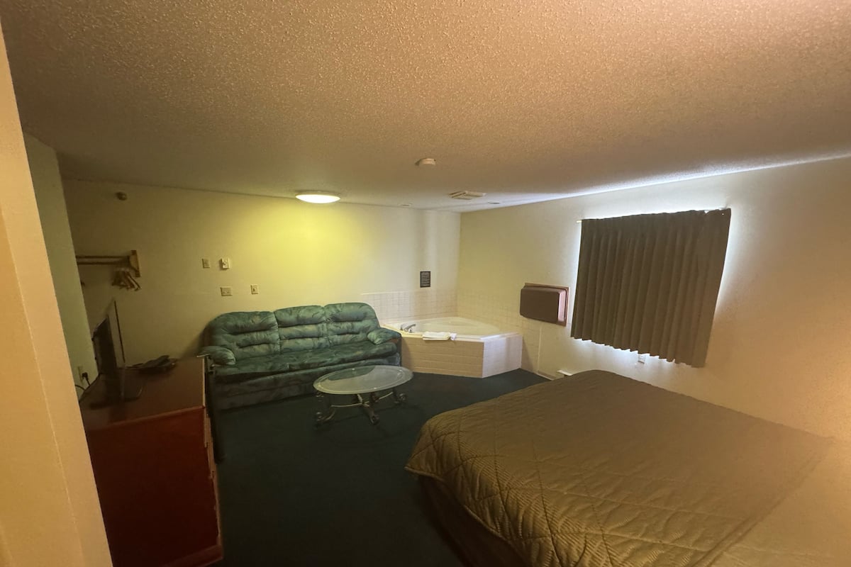 A compact hotel room with a single bed, a green sofa, a small table, a bathtub, and a desk.