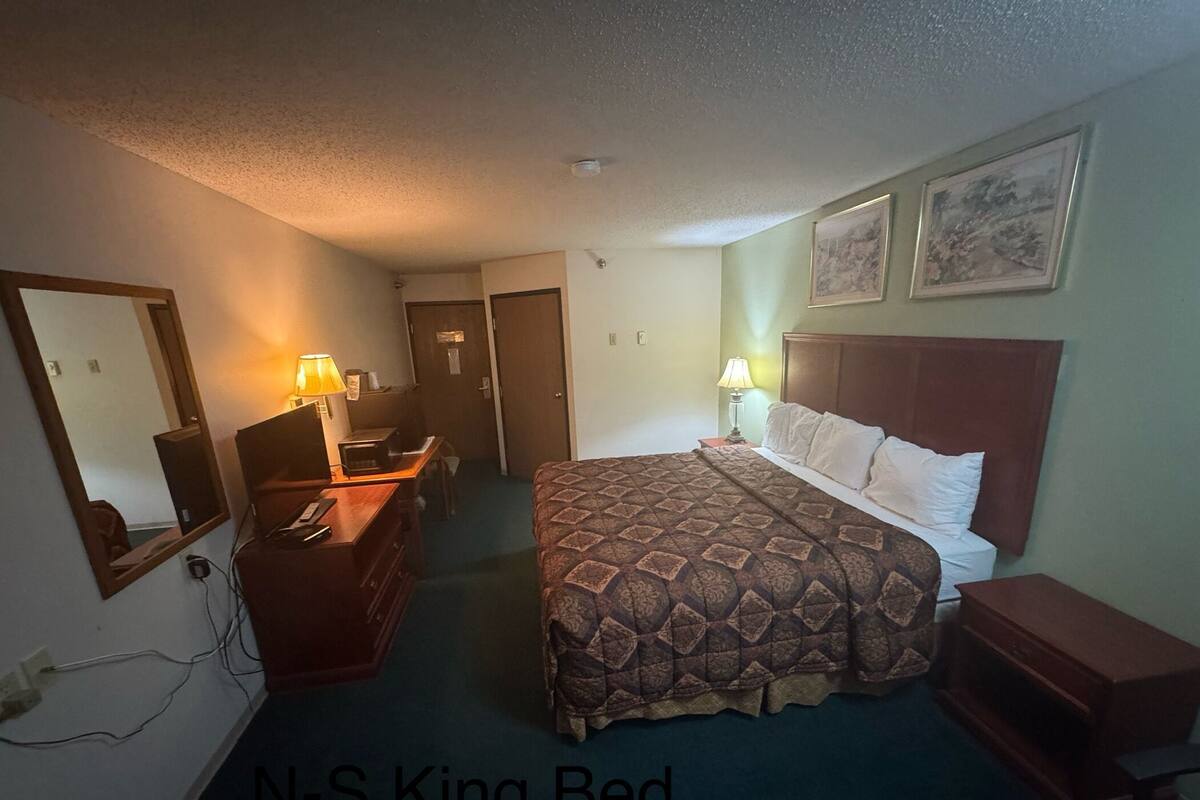 A hotel room with a king bed, a desk with a lamp, a mirror, and a door leading to another room.