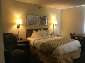 A hotel room with a bed, two bedside lamps, a chair, a table, and two framed pictures on the wall. - Smart Choice Inn  (Plankinton)