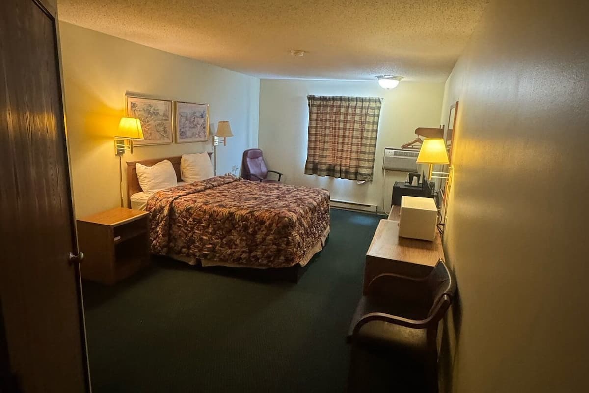 A hotel room with a bed, a desk, a chair, a lamp, and a trash bin.