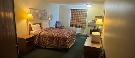 A hotel room with a bed, two bedside tables, a desk, a chair, a lamp, a window with curtains, and a wall-mounted air conditioning unit.