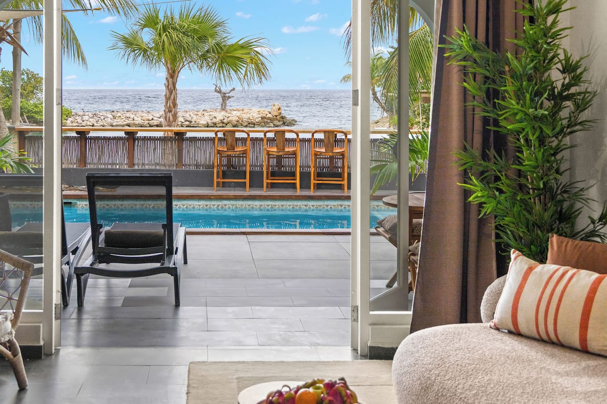 A room with a view of the ocean, a pool, and a seating area with chairs.