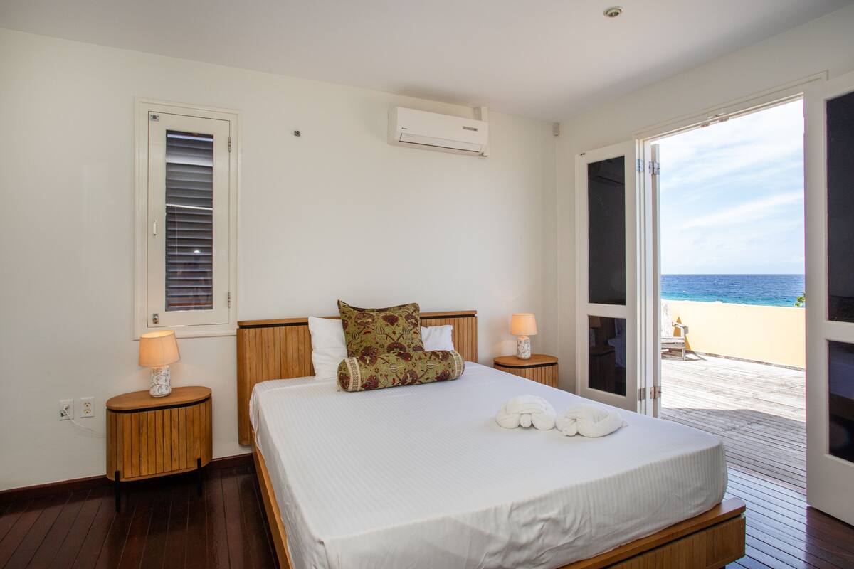 A bedroom with a bed, two bedside tables, a window with blinds, an air conditioning unit, and a view of the beach through an open door.