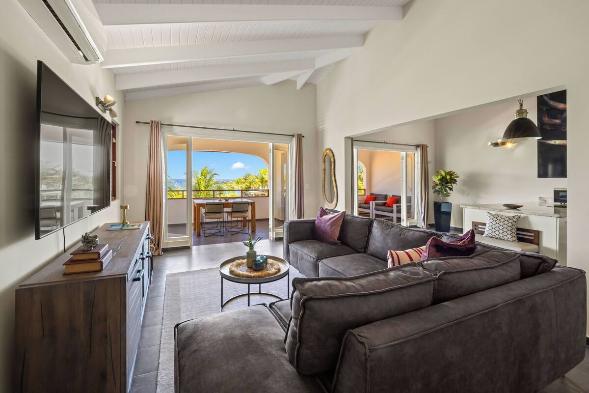 A modern living room with a sofa, a coffee table, and a TV. There is a balcony with a view of palm trees.