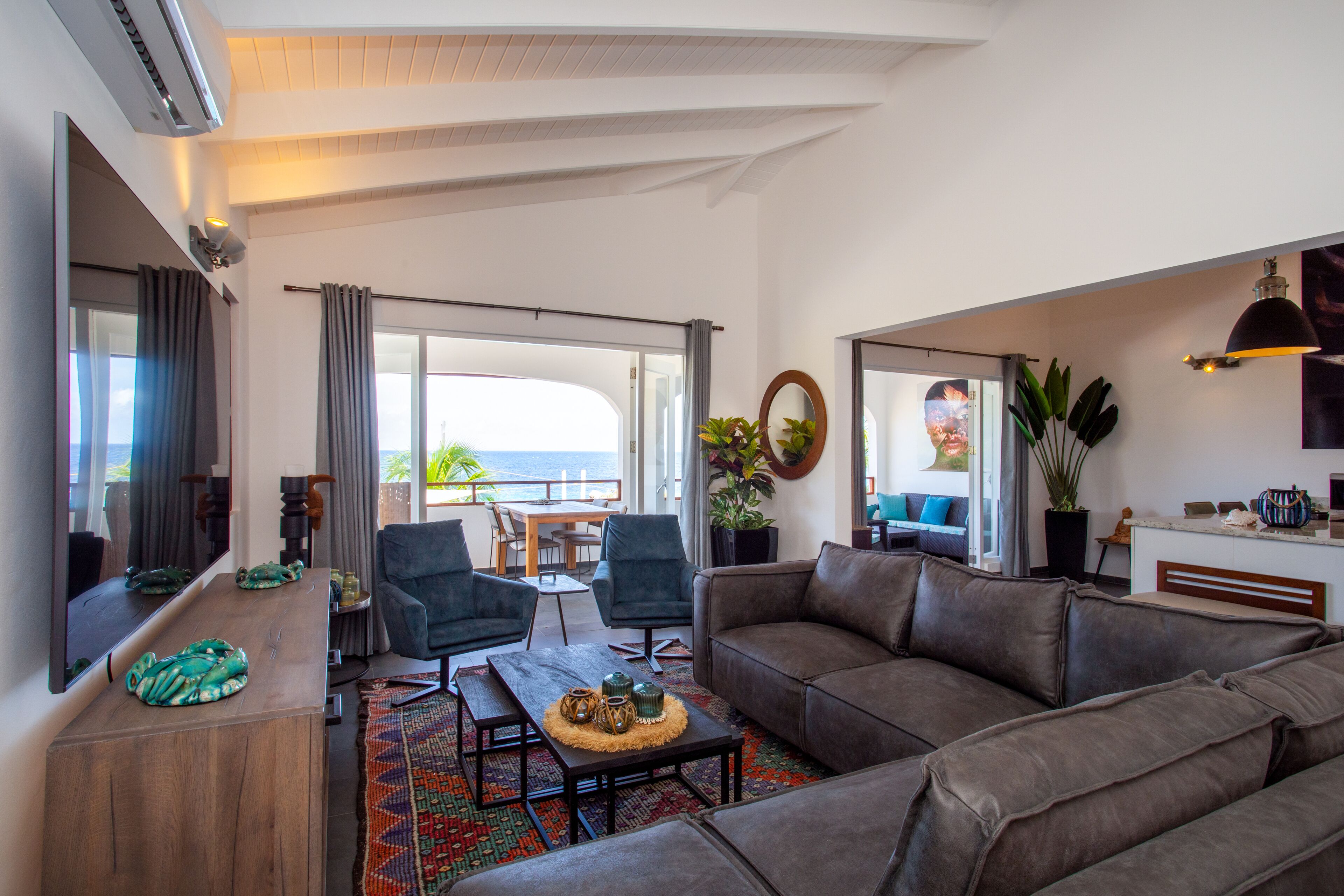 A spacious living area with a large sofa, a coffee table, and a dining table. There is a large window offering a view of the ocean.