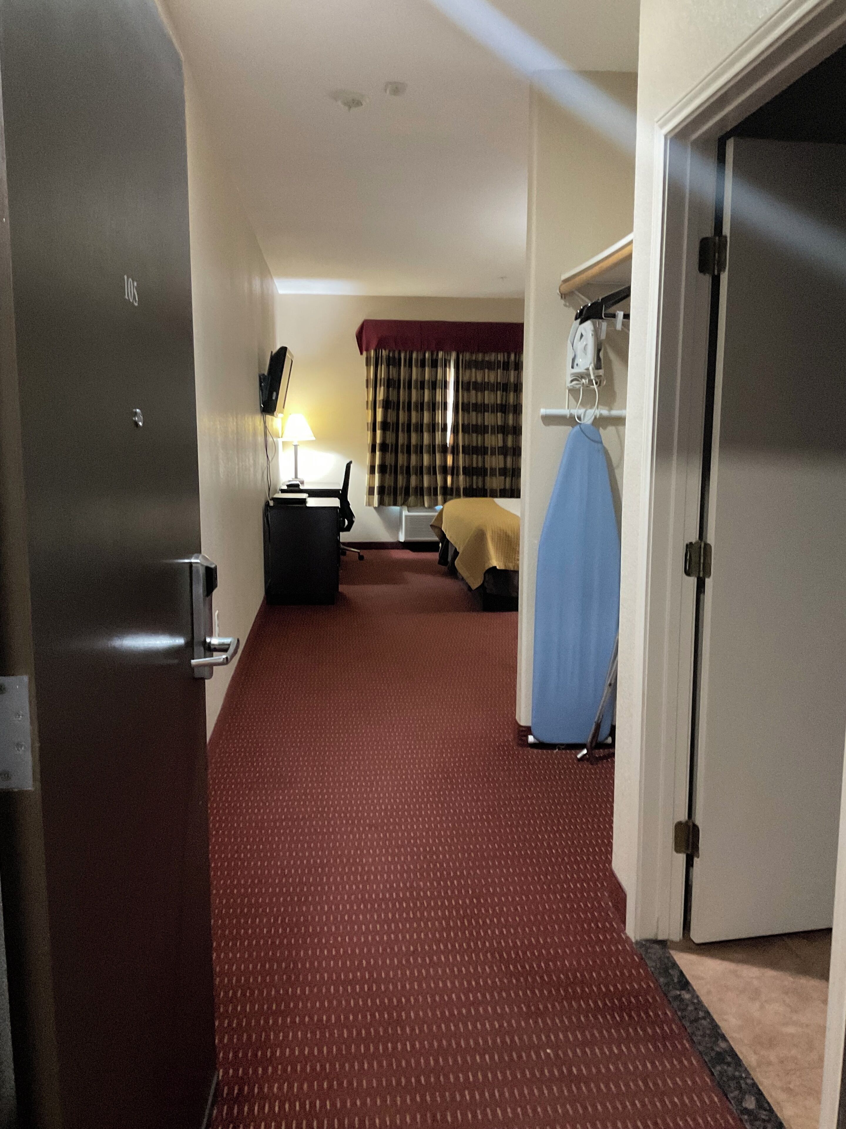 A hotel room with a red carpet, a bed, a desk with a lamp, a window with curtains, and a door.