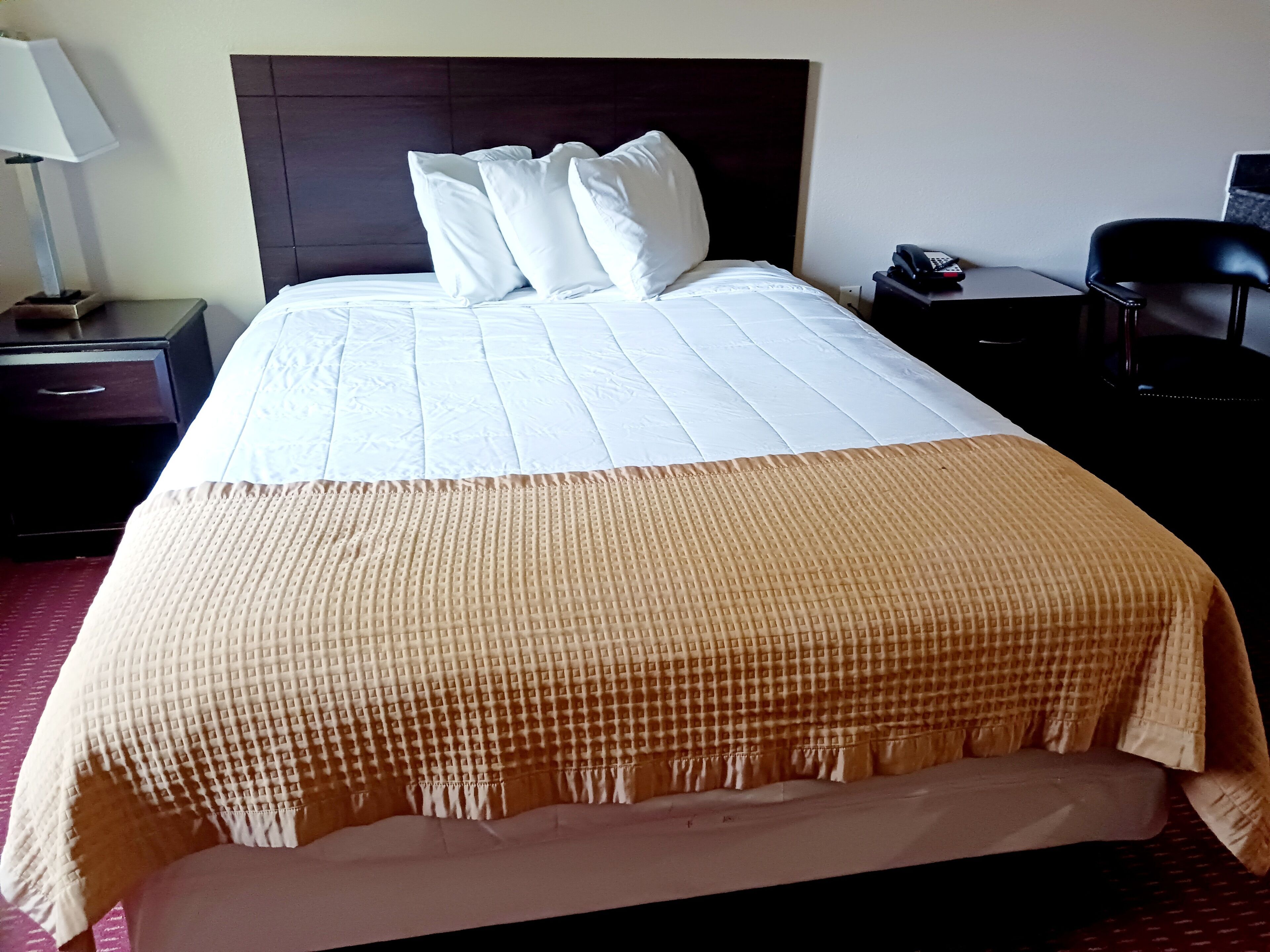 Een net opgemaakt bed met witte lakens en een bruine decoratieve deken, een nachtkastje met een lamp, en een stoel.