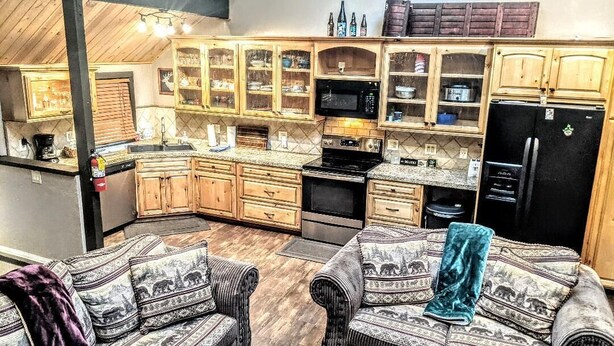 A cozy kitchen with wooden cabinets, a microwave, and a refrigerator. There are two sofas with patterned cushions and a blanket. - Escape For All Seasons (Big Bear Lake)