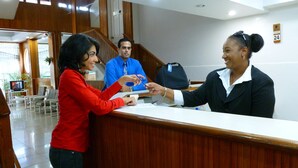 A reception area with a wooden reception desk, a TV on a stand, and a staircase in the background. - Royal Palm Suite Hotel (Port of Spain)