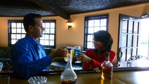 A man and a woman sitting at a bar with drinks, a bottle of liquor, and a glass carafe. - Royal Palm Suite Hotel (Port of Spain)