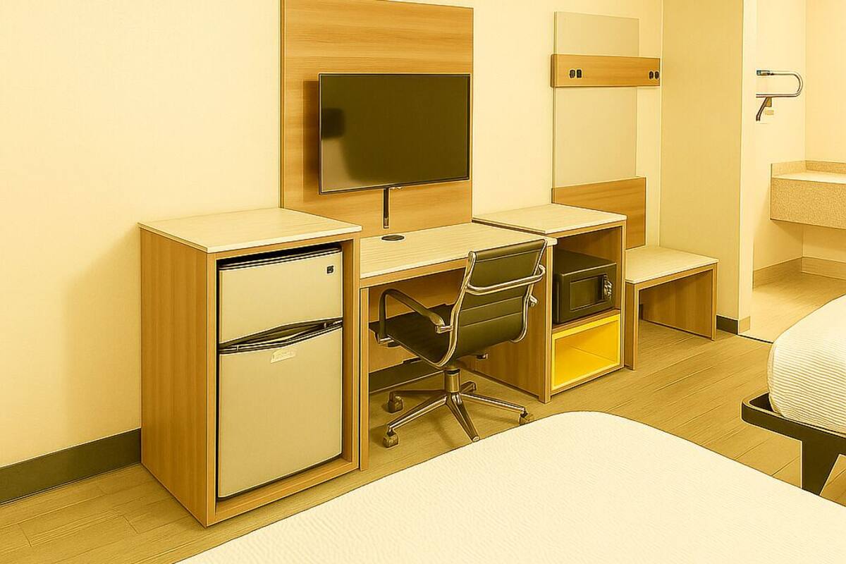 A hotel room with a bed, a desk with a chair, a television, a mirror, a small refrigerator, and a bathroom with a sink and toilet.