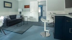 A compact hotel room with a dark blue sofa, a small round table, a white chair, and a kitchenette area. - Atlantic Inn (Wethersfield)