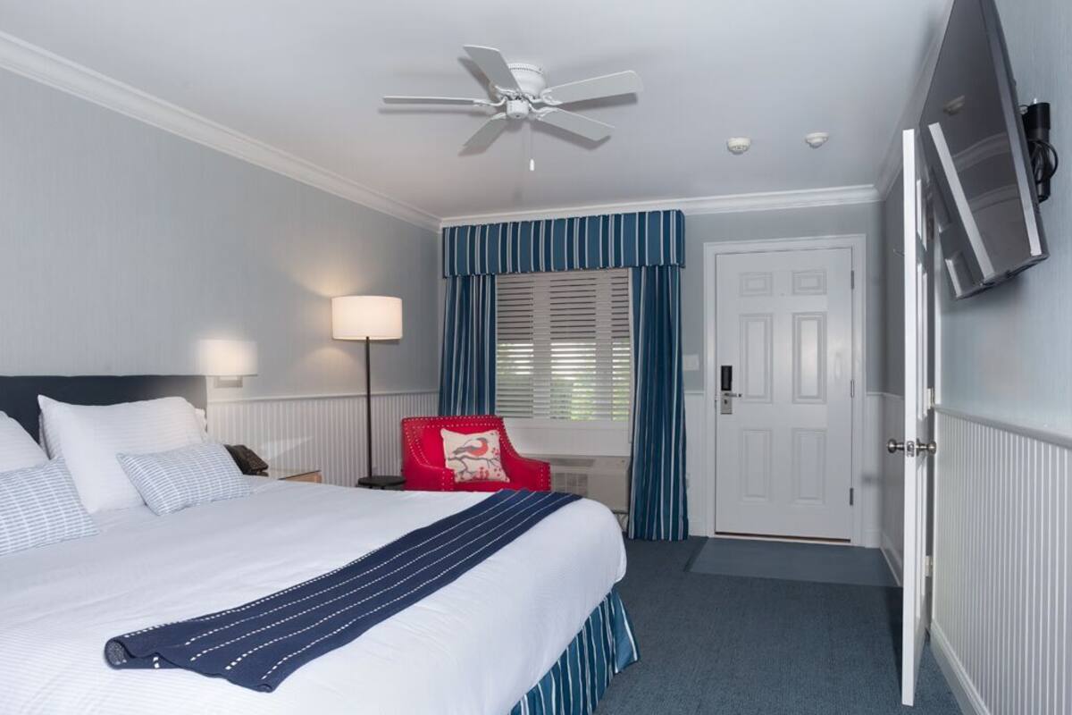 A hotel room with a bed, a ceiling fan, a TV, a red chair, and a door.
