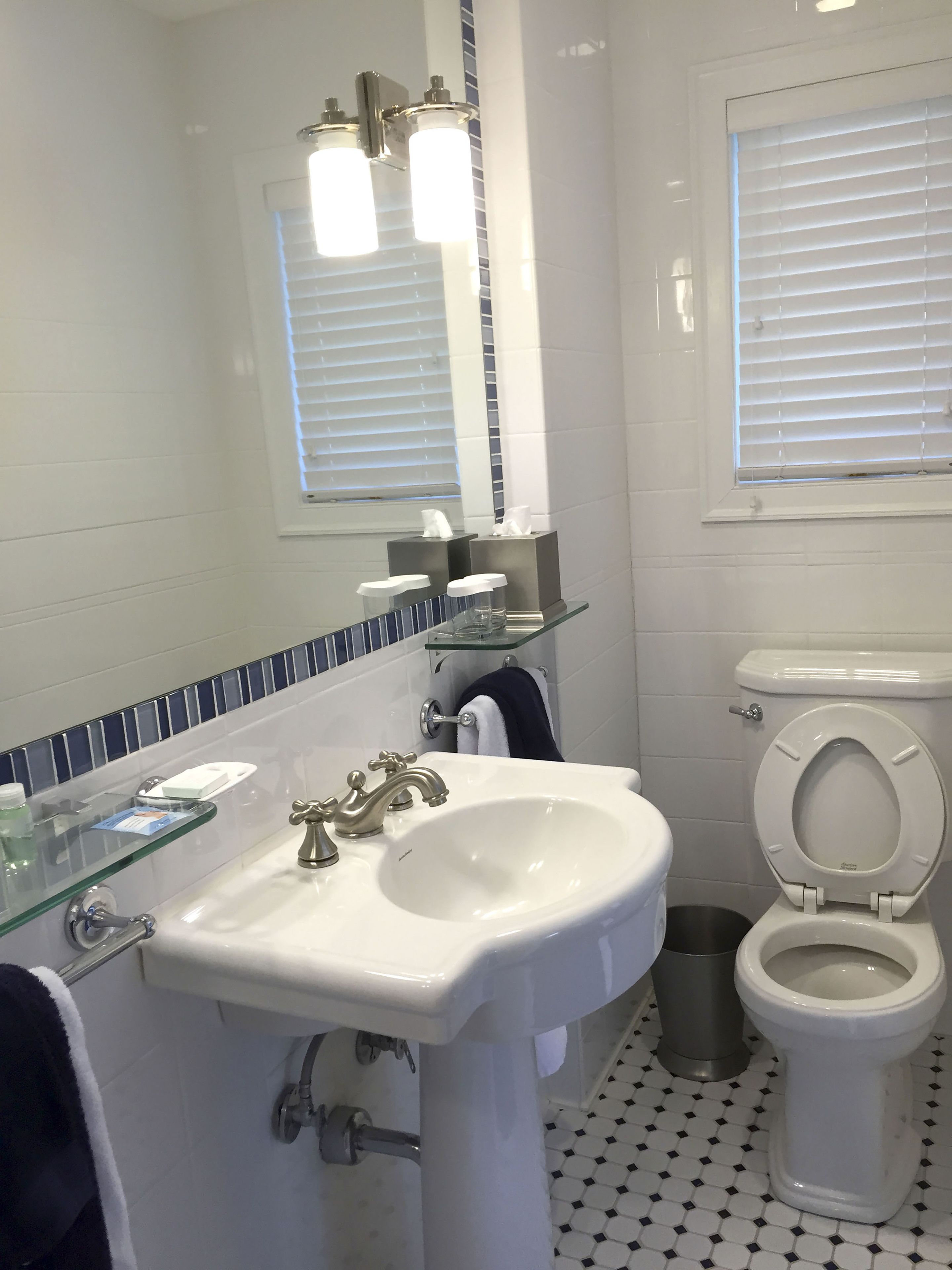A bathroom with a white sink, two faucets, a mirror, a toilet, and a window with blinds.