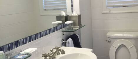 Un baño con lavamanos blanco, dos llaves, un espejo, inodoro y ventana con persianas.