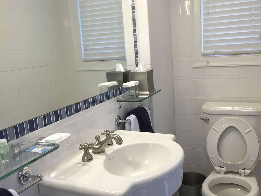 A bathroom with a white sink, two faucets, a mirror, a toilet, and a window with blinds.