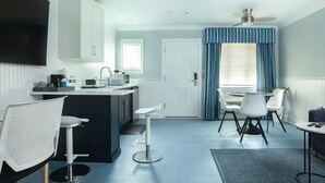 A modern kitchen with white cabinets, a central island, and bar stools. There is a dining area with a table and chairs, and a large window with blue curtains. - Atlantic Inn (Wethersfield)