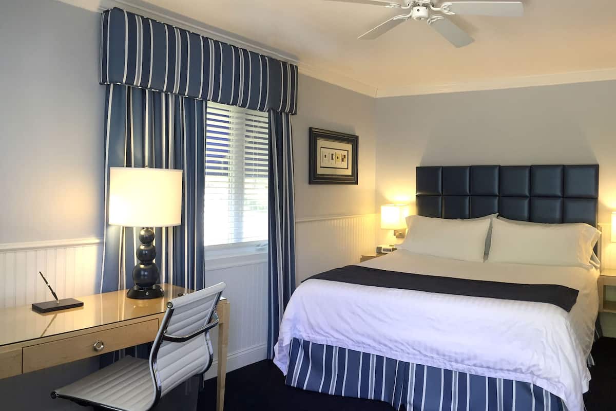 A hotel room with a bed, bedside lamps, a desk with a lamp, a chair, and a ceiling fan.