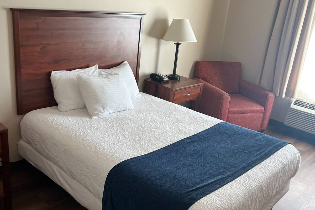 A hotel room with a large bed, a wooden headboard, a nightstand with a lamp, a red chair, and a window with curtains.