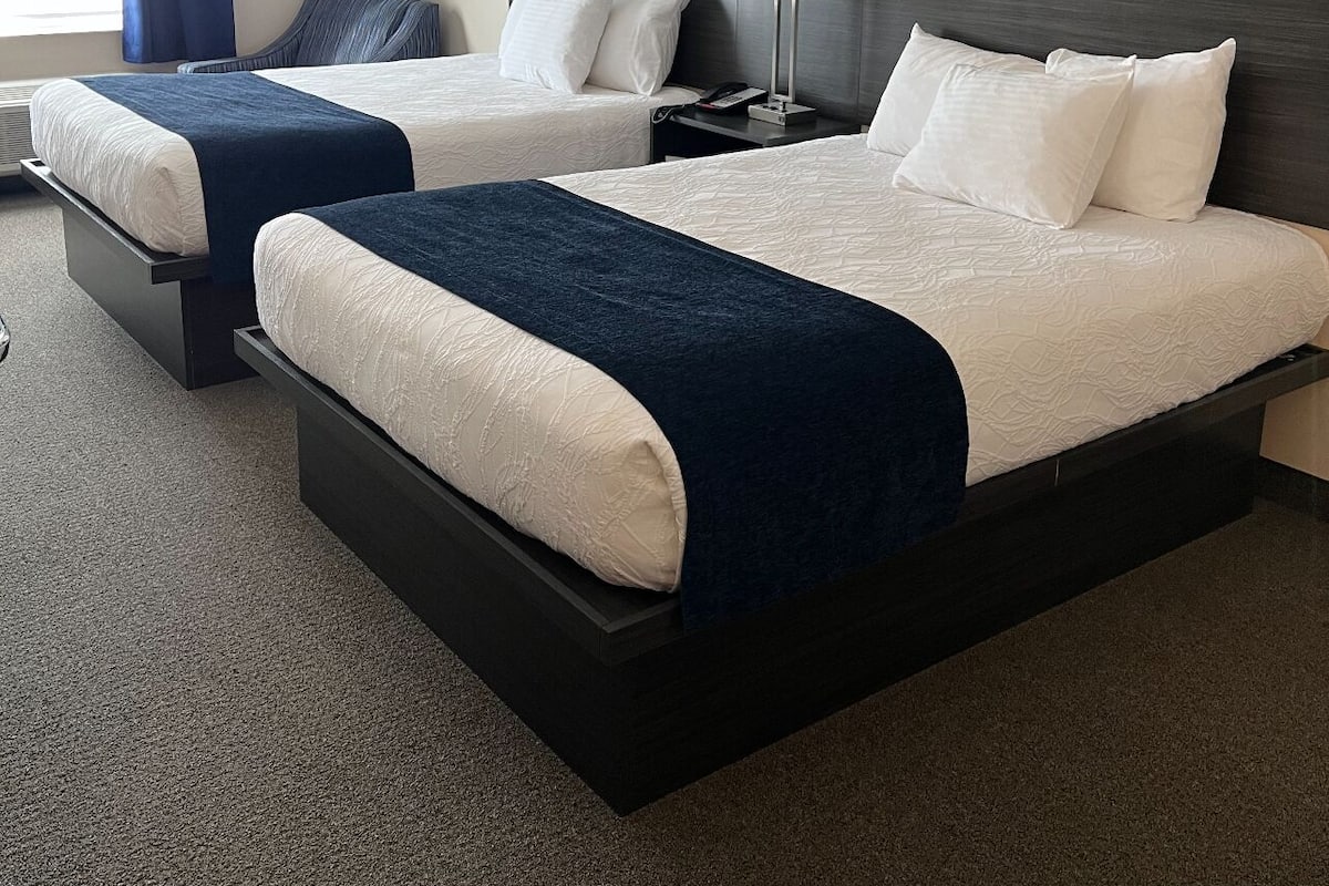 A hotel room with two beds, a dark headboard, a bedside table with a lamp, and a window with curtains.