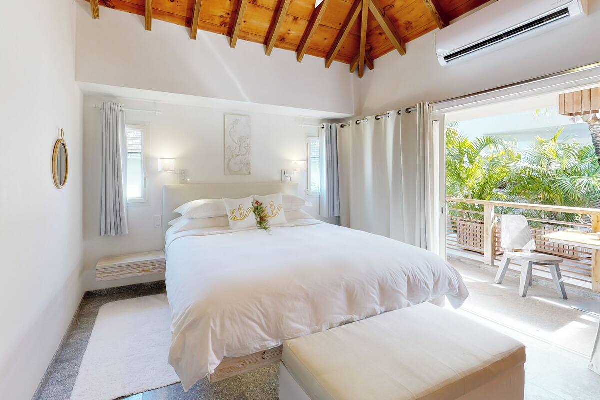 Ein Schlafzimmer mit einem großen Bett, einer Bank, einem Balkon mit Sitzmöbeln und Blick auf Grünflächen.