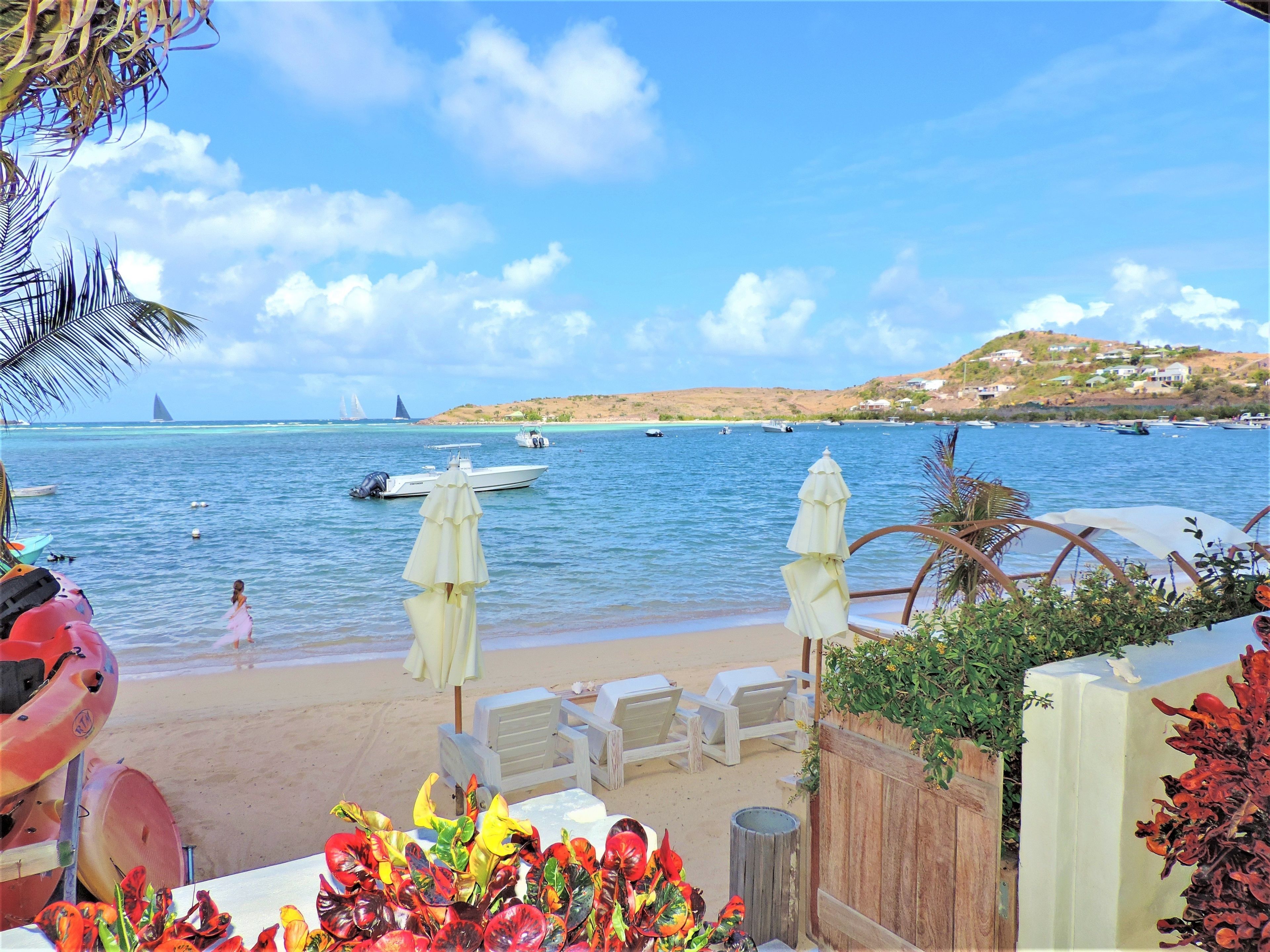 A beachside view with a sandy area, white umbrellas, and lounge chairs. Boats are visible on the water, and there is a hill with buildings in the background.