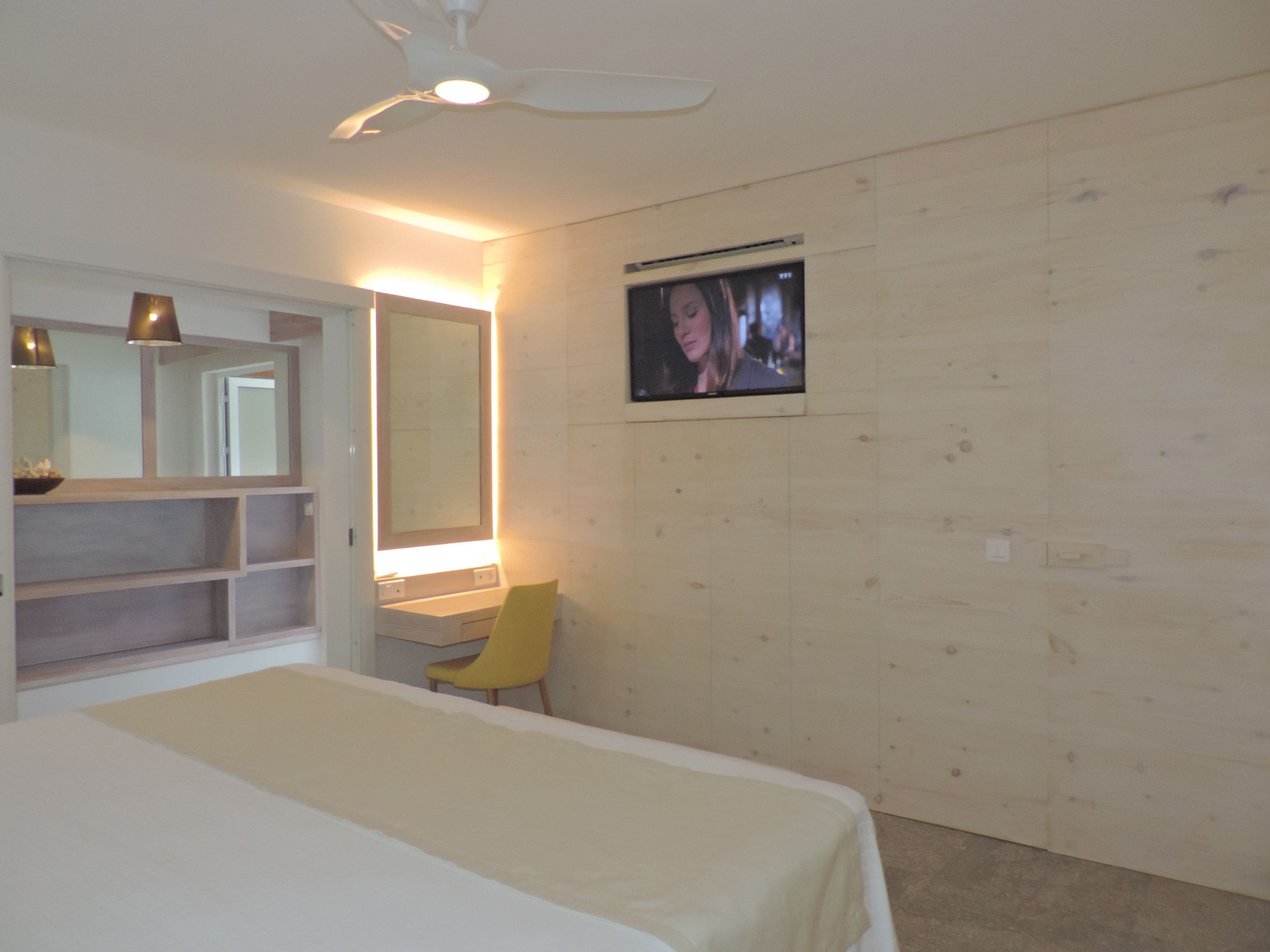 A bedroom with a bed, a chair, a TV mounted on the wall, and a ceiling fan.