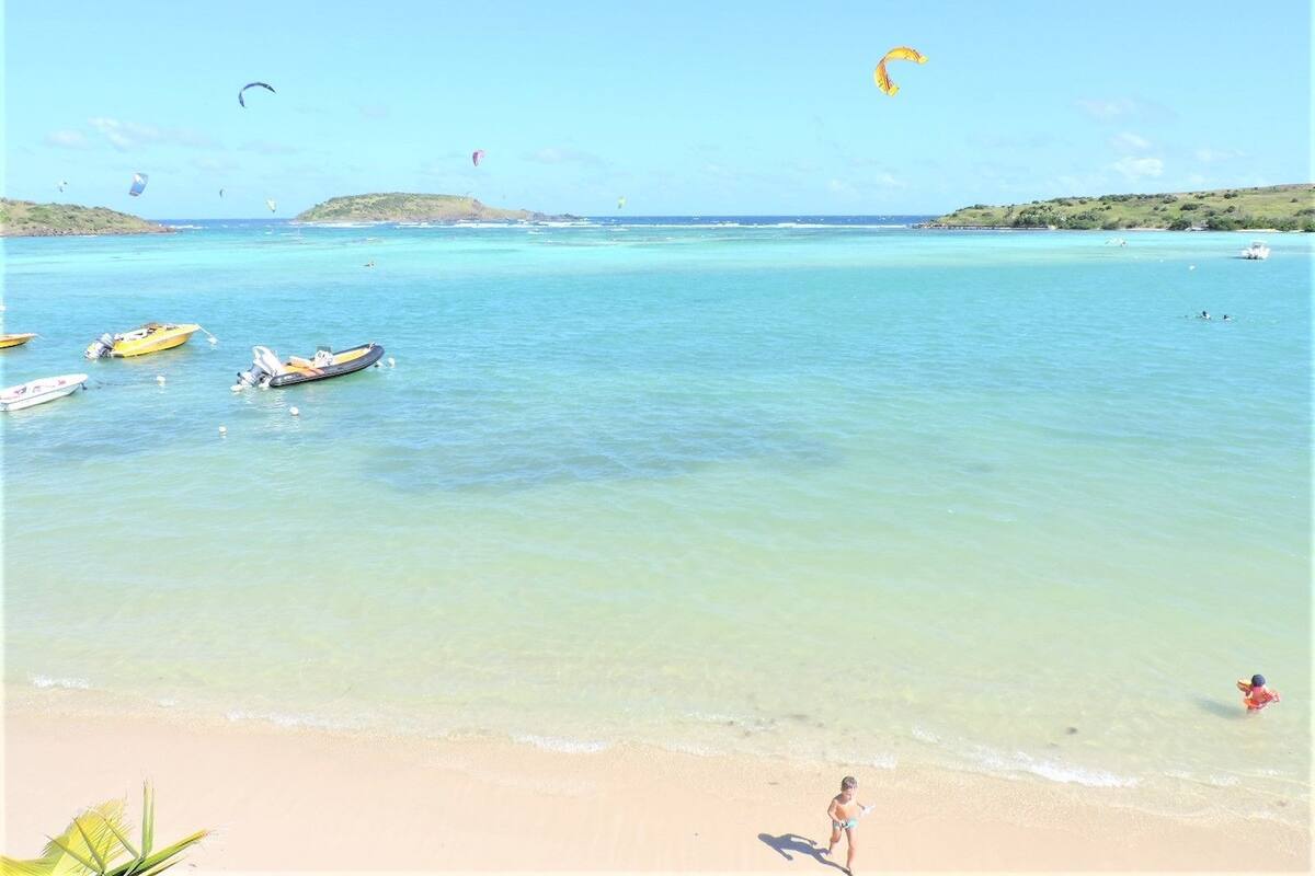 Ein Strand mit klarem türkisblauem Wasser, mehreren Booten und einer Person, die am Ufer entlanggeht.
