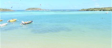 A beach with clear turquoise water, several boats, and a person walking along the shore.