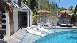 Zona de piscina con sillas de descanso, una fuente de agua y un pequeño edificio con una puerta.