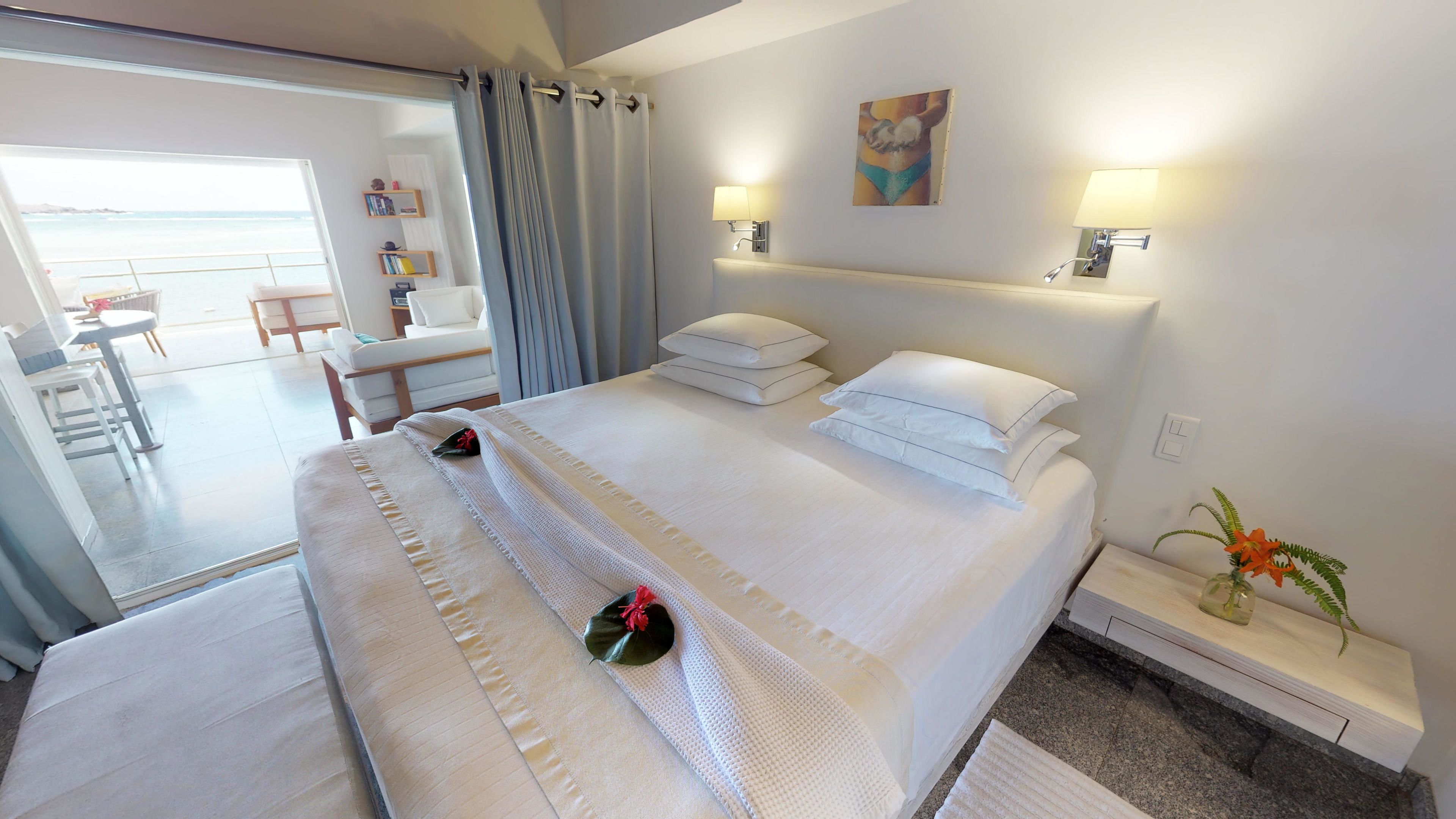 A hotel room with a large bed, two bedside tables, a small desk, a chair, a view of the sea, and a painting on the wall.