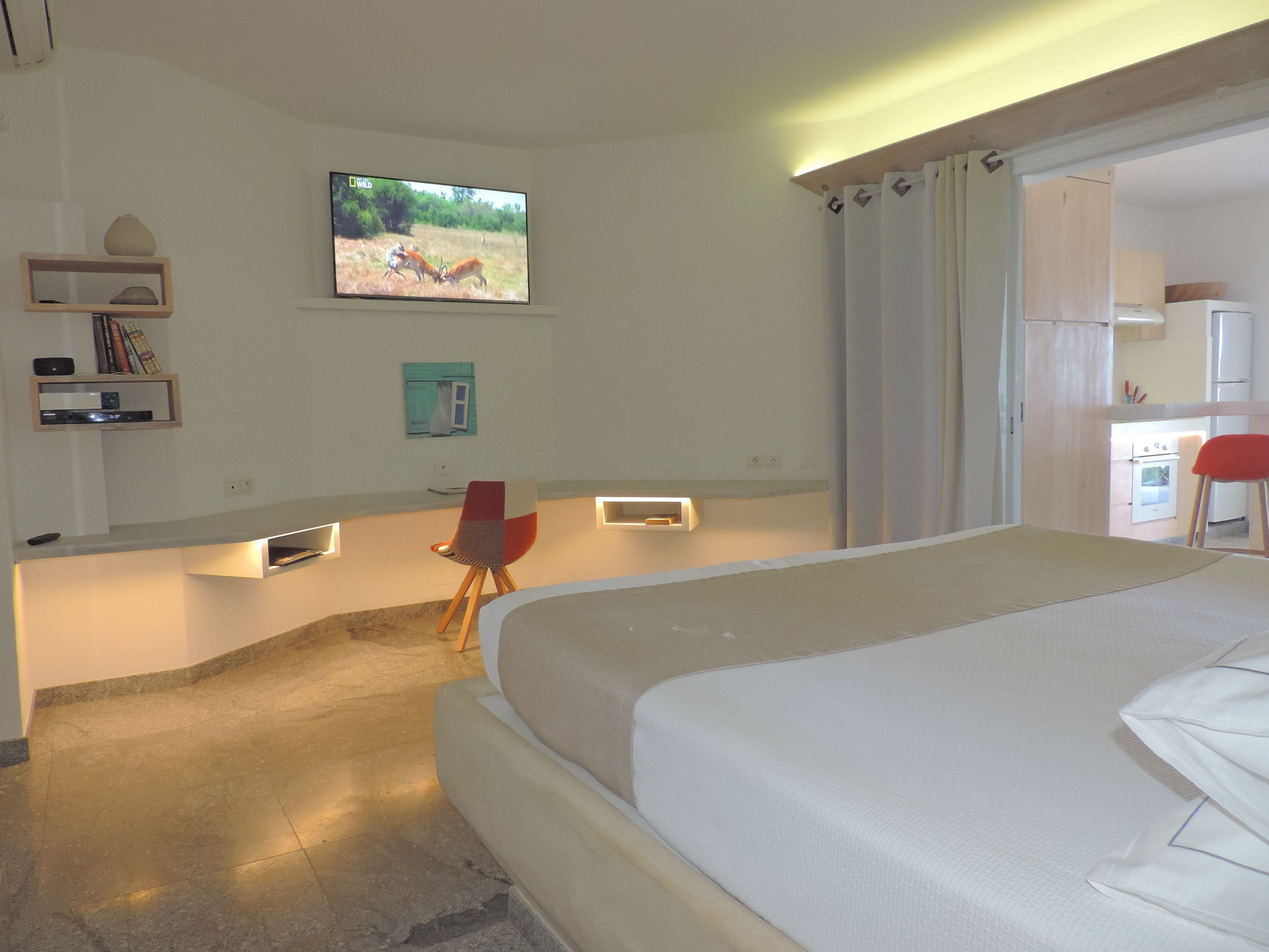 A modern hotel room with a large bed, a desk with a computer, a TV mounted on the wall, and a small kitchenette.