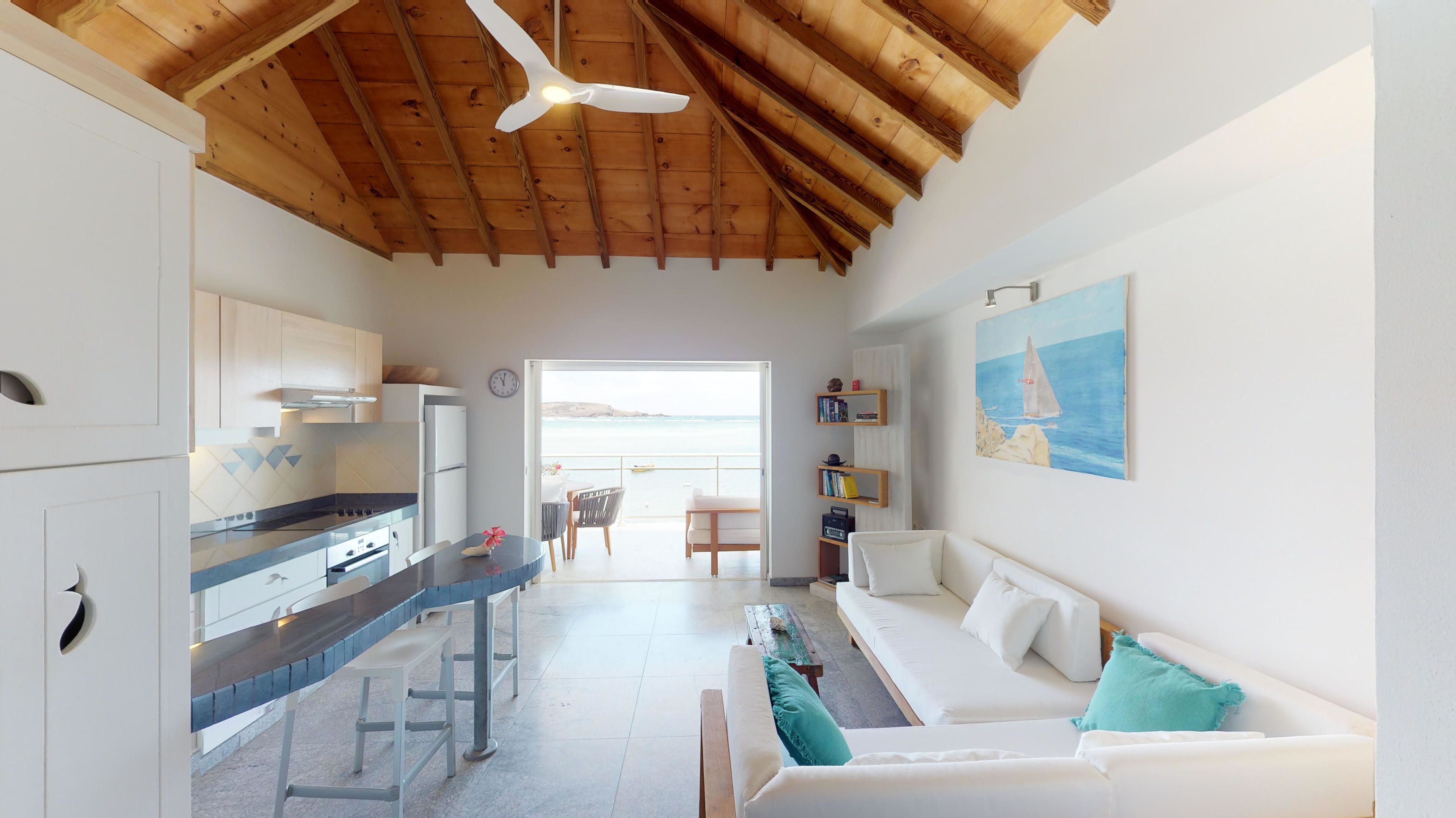 Una cocina moderna con armarios blancos y techo de madera, un área de comedor con un sofá blanco y un cuadro de un velero, y vista al mar a través de una puerta abierta.