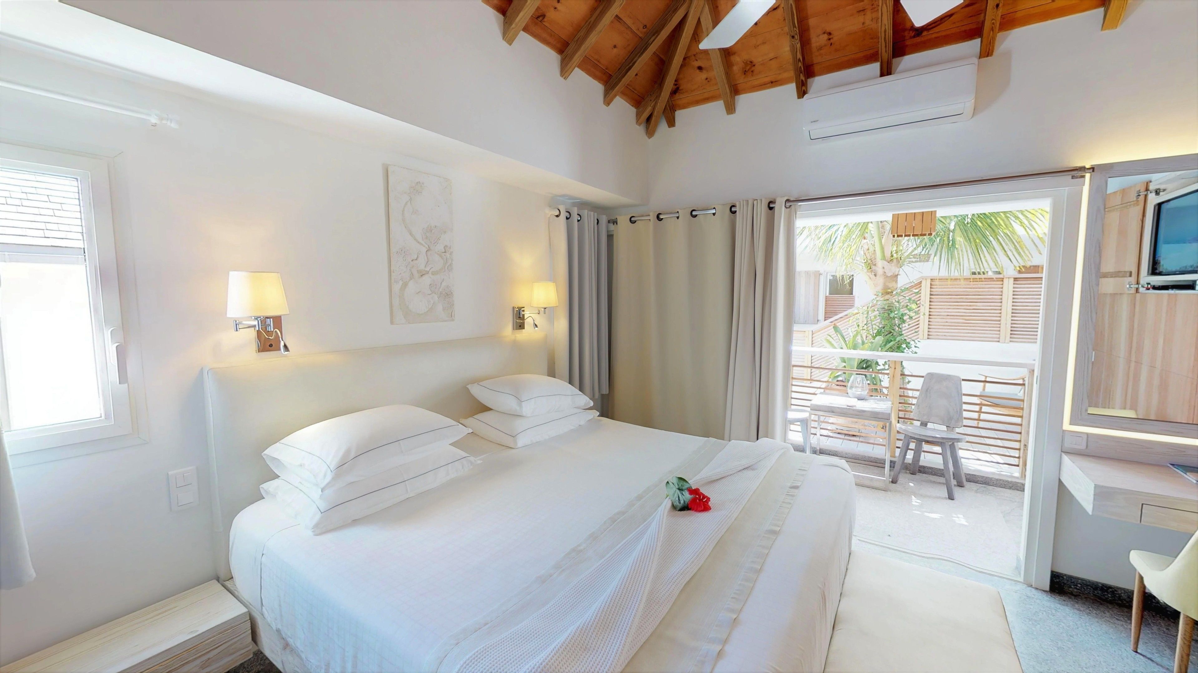 A hotel room with a large bed, two bedside lamps, a ceiling fan, and a sliding glass door leading to an outdoor area with a table and chairs.