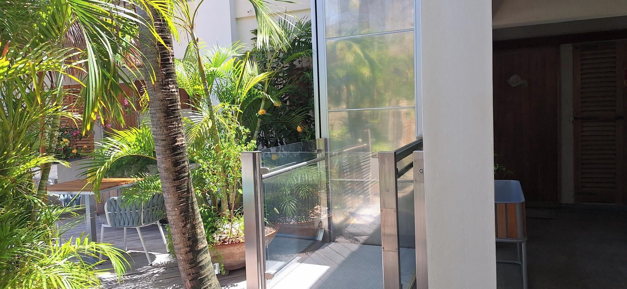 Un edificio moderno con balcón de vidrio que da a un jardín con palmeras y otra vegetación.