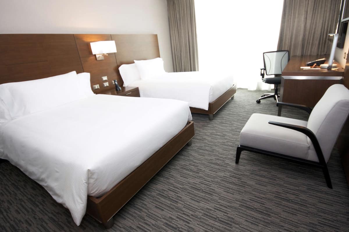 A hotel room with two beds, a desk, a chair, and a lamp.