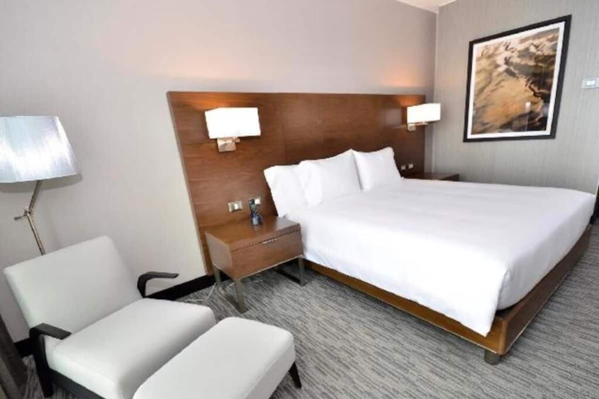 A hotel room with a large bed, two white chairs, a nightstand, and a lamp.