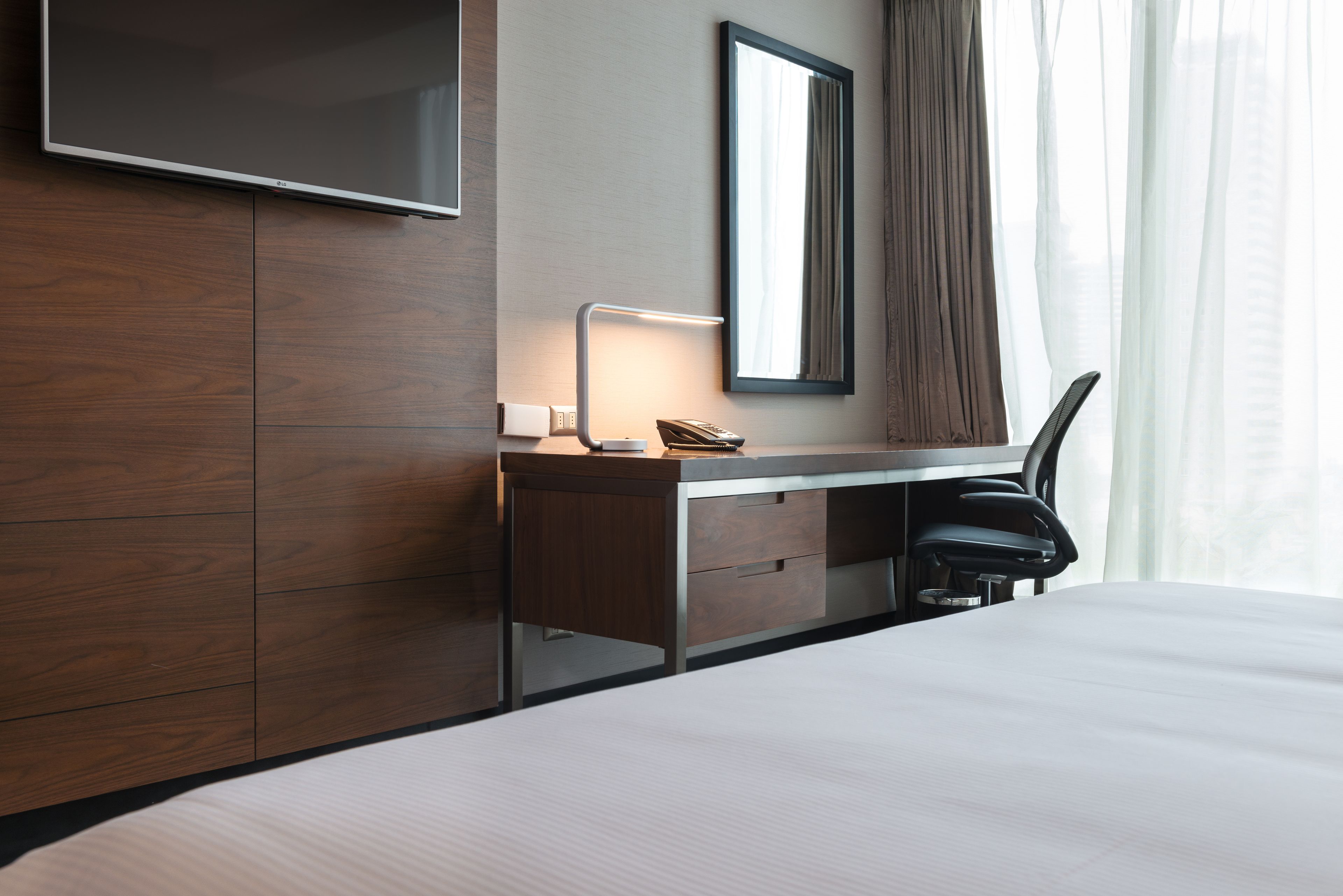 A hotel room with a wooden desk, a flat-screen TV, a mirror, a telephone, and a chair.