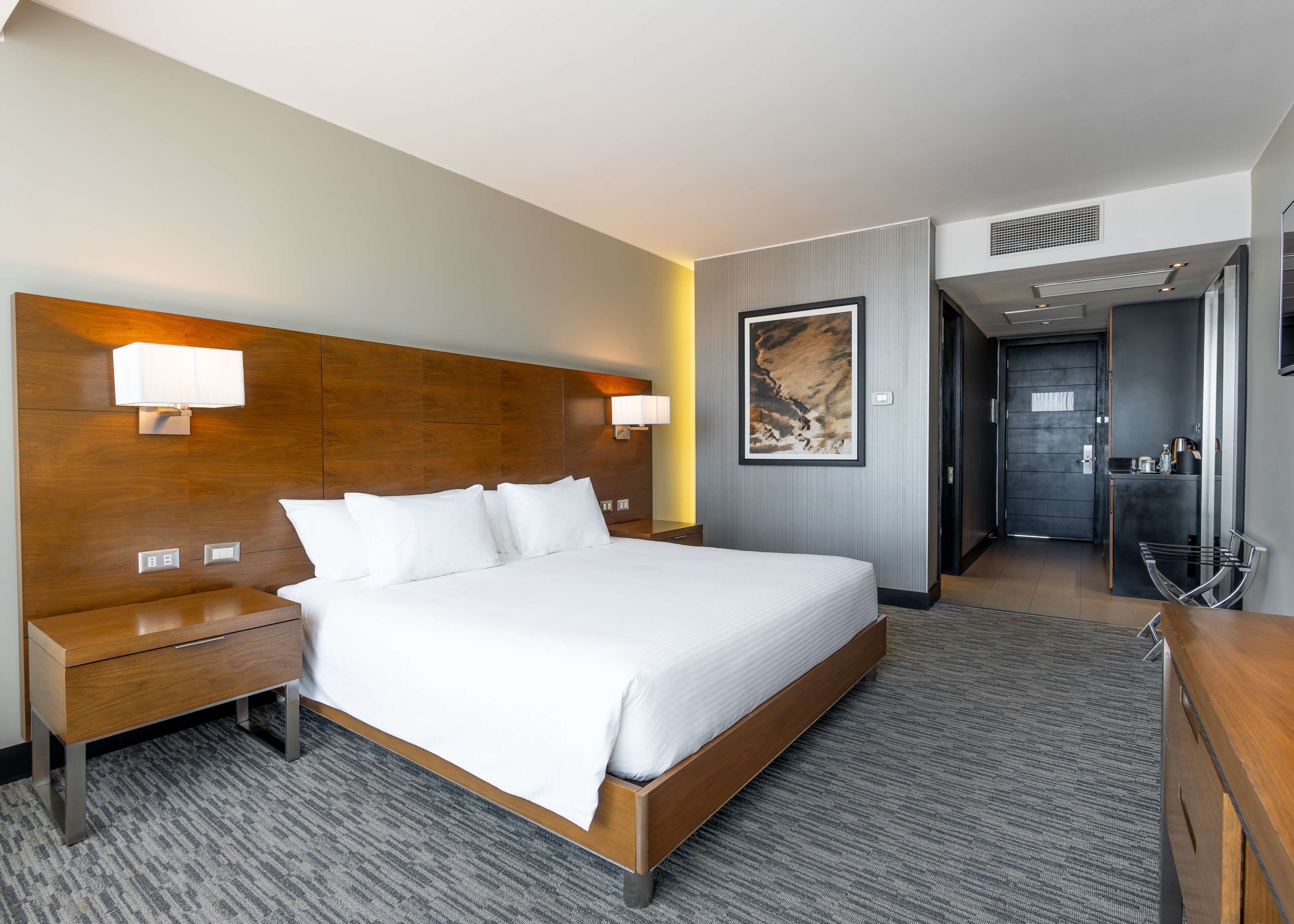 Una habitación de hotel moderna con una cama grande, cabecera de madera, lámparas de noche, una obra de arte enmarcada y un baño visible a través de una puerta abierta.