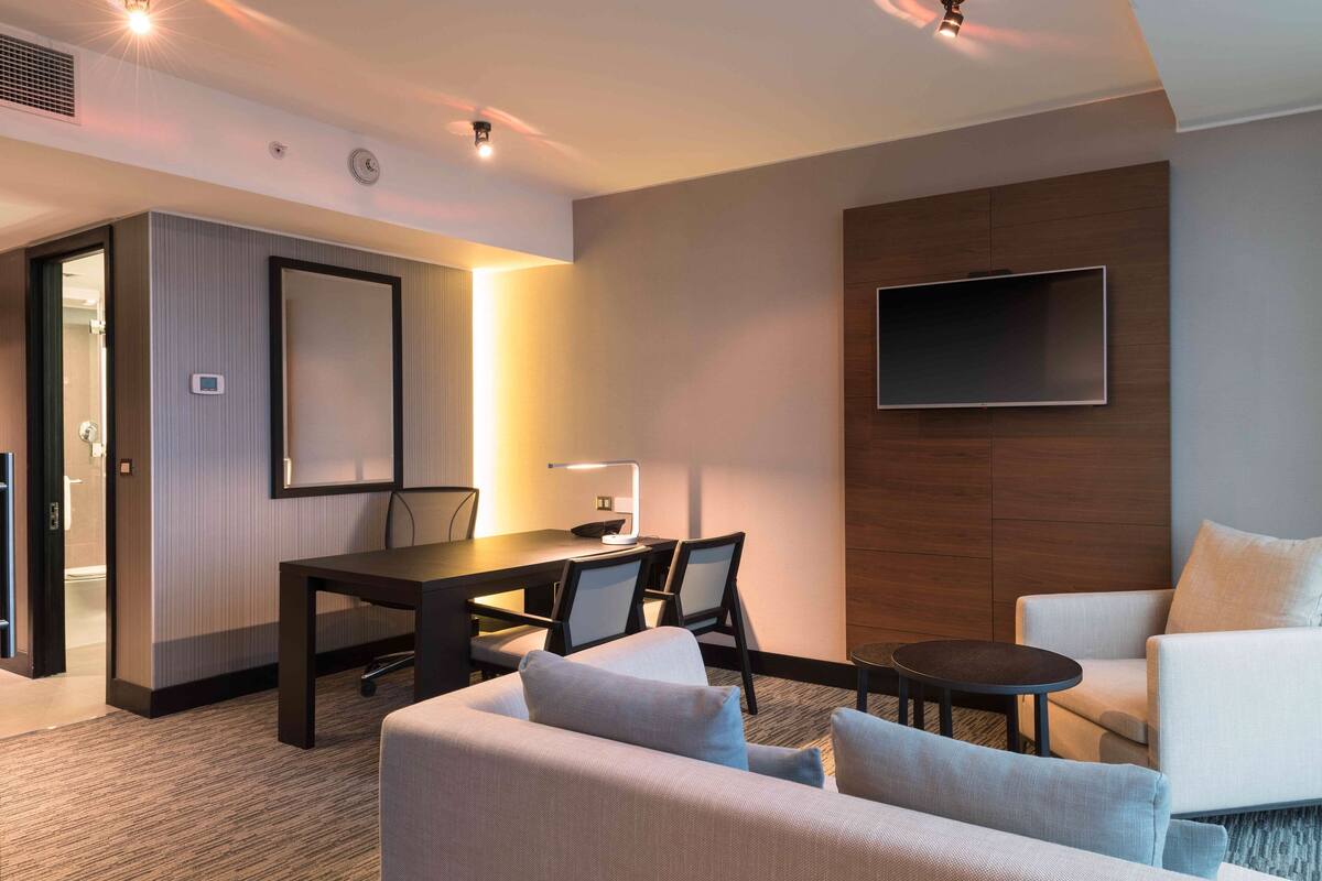 A modern hotel room with a sofa, a dining table with chairs, a TV mounted on the wall, and a small round table.
