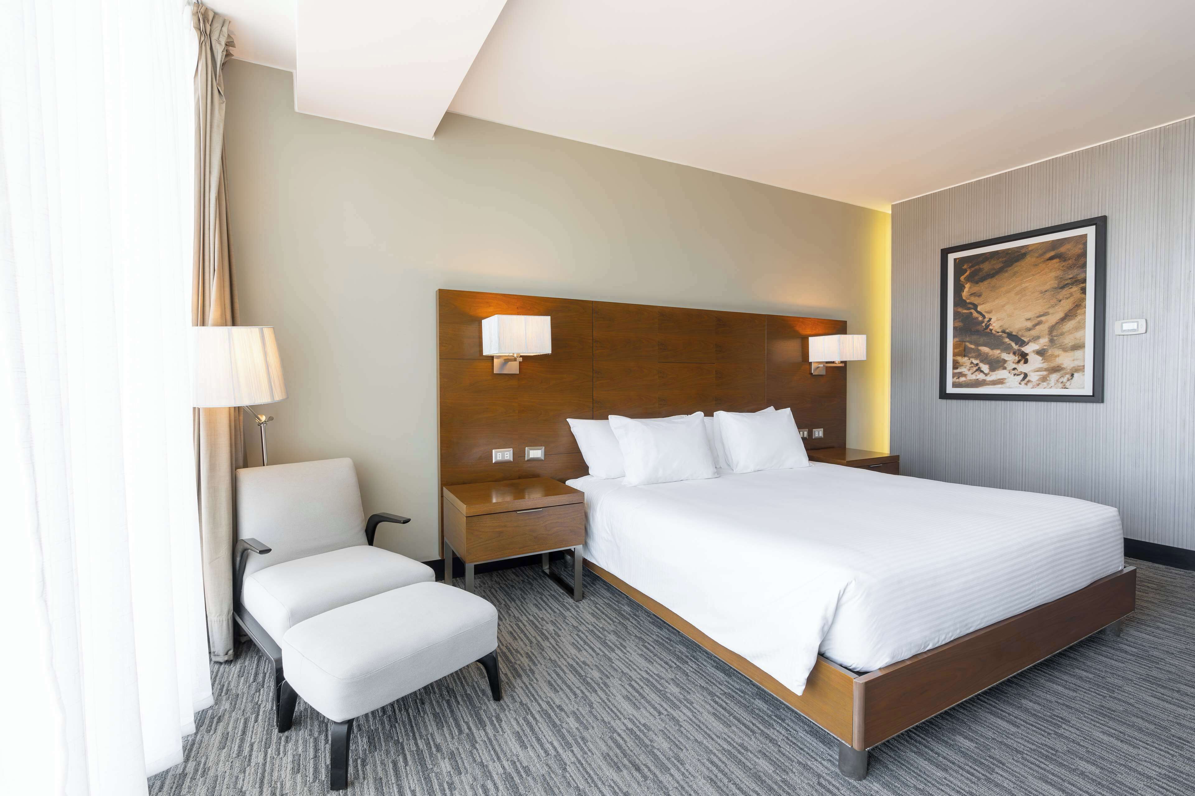 Habitación de hotel con una cama grande, una mesita de noche, una silla y una lámpara de pared.