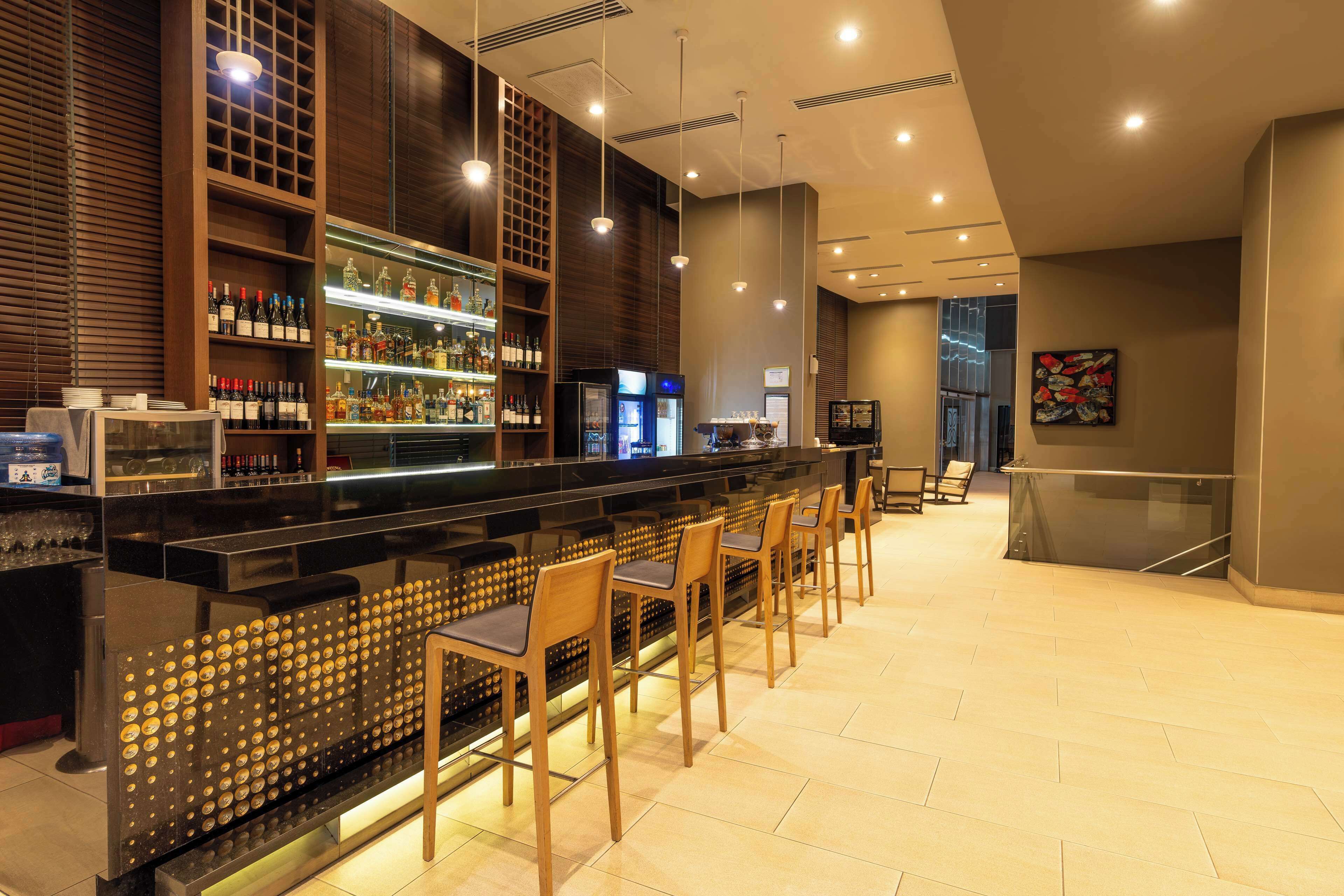 Una zona de bar con una barra larga, varios estantes llenos de botellas y múltiples taburetes.