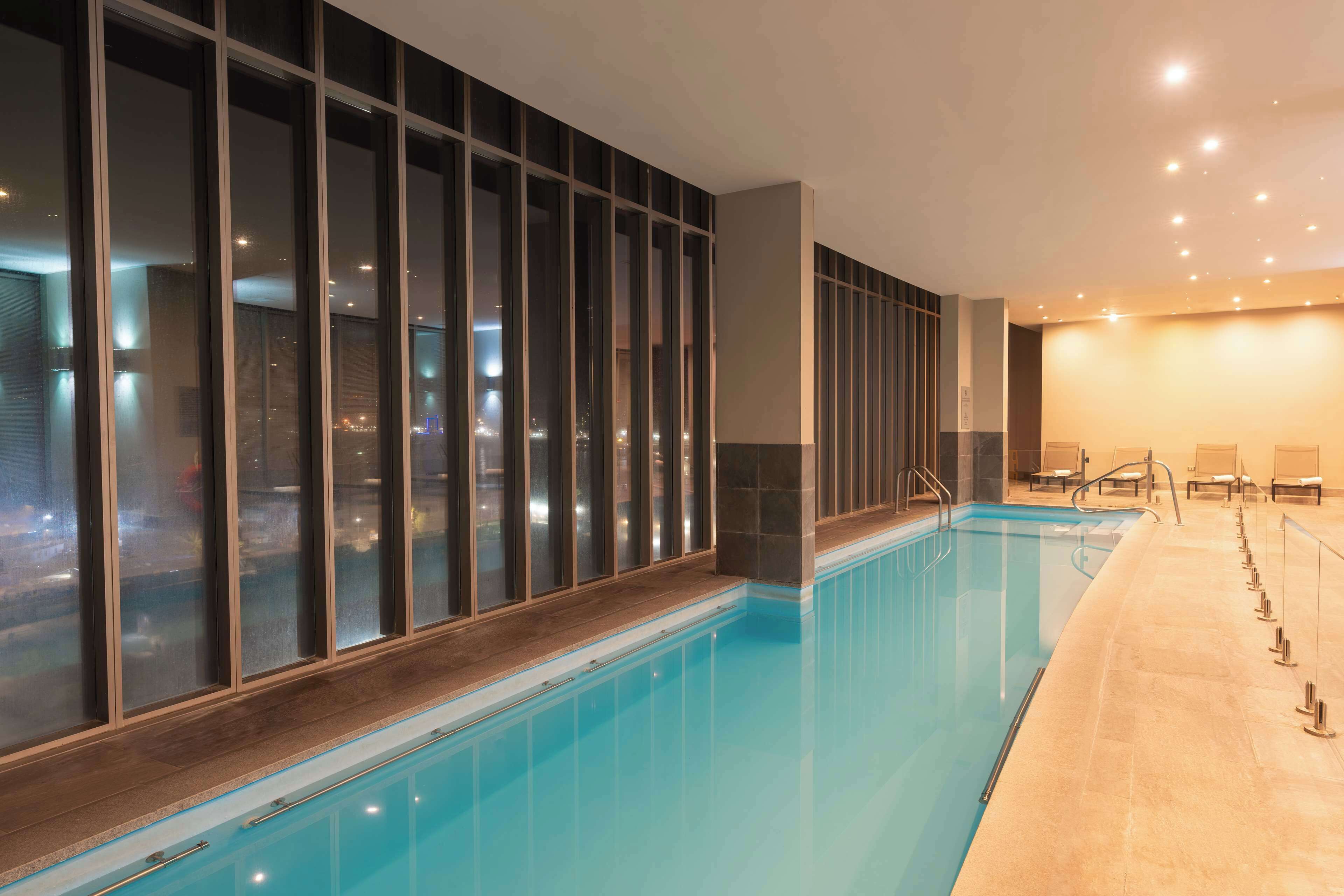 An indoor swimming pool with clear glass walls, a tiled floor, and a seating area with chairs and a table.