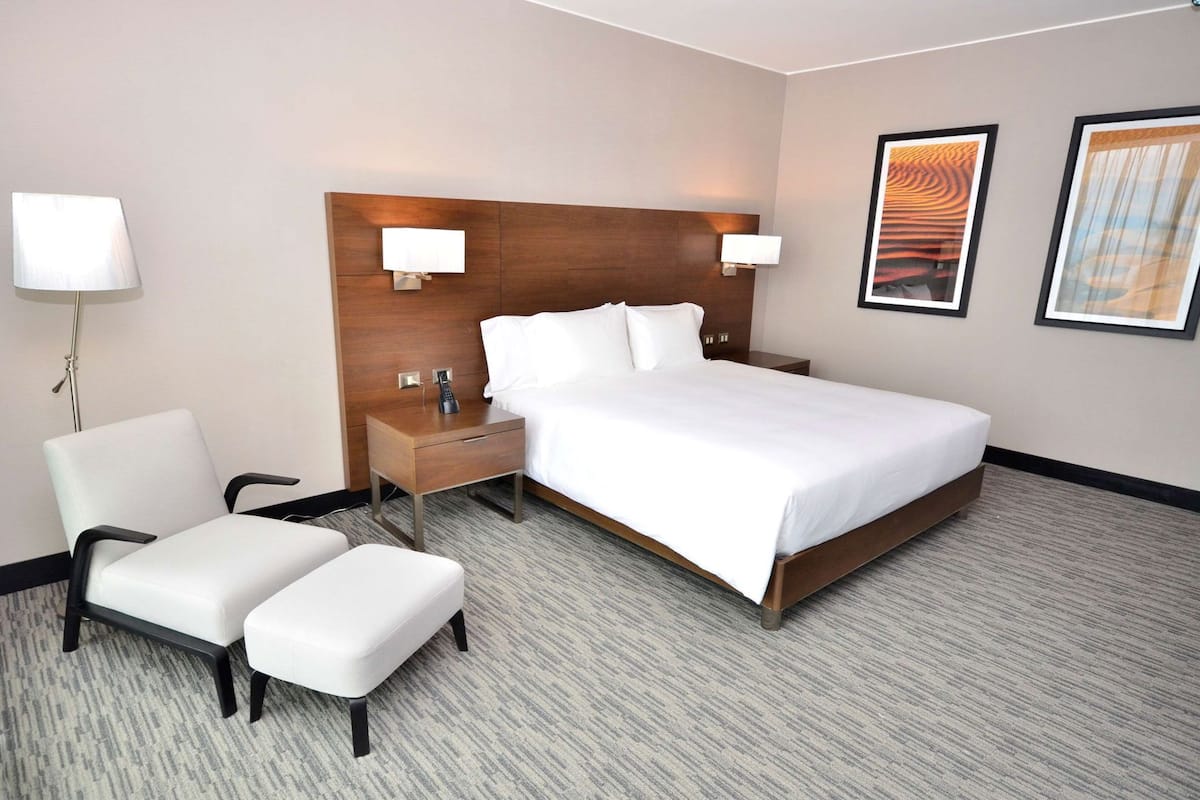 A hotel room with a large bed, a bedside table with a lamp, a chair, and two framed pictures on the wall.