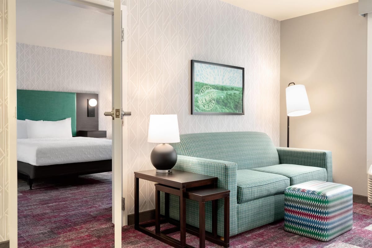 Une chambre d’hôtel avec un lit, un canapé vert, une petite table avec une lampe, un tableau au mur et un pouf à motifs.