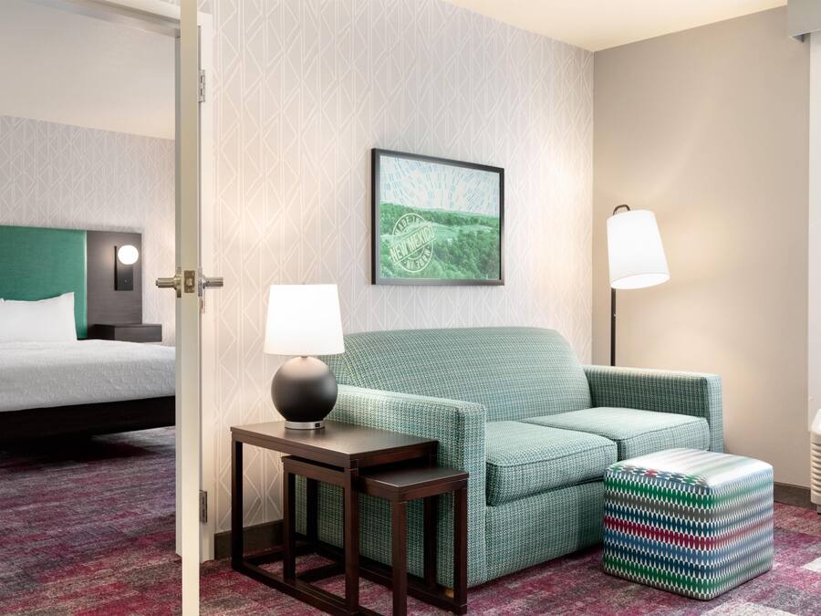 Ein Hotelzimmer mit einem Bett, einem grünen Sofa, einem kleinen Tisch mit Lampe, einem Bild an der Wand und einem gemusterten Hocker.