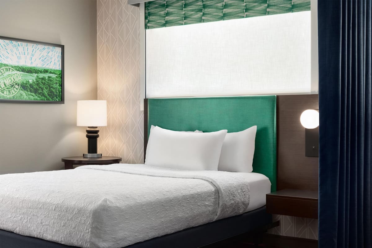 Une chambre d’hôtel avec un lit, une table de chevet avec une lampe, une tête de lit verte et un luminaire fixé au mur.