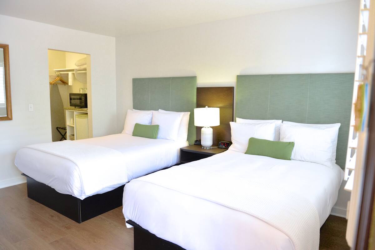 A hotel room with two beds, each with white bedding and green pillows, a bedside table with a lamp, and a small TV.